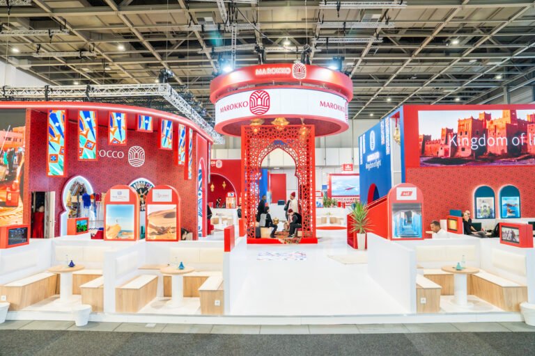 L’ONMT donne une nouvelle dimension au Pavillon Maroc sur les grands rendez-vous mondiaux du Tourisme Vue d'ensemble du Pavillon Maroc sur un salon professionnel avec ses espaces de réunion, sa structure en arche rouge ciselée et ses affiches promotionnelles.