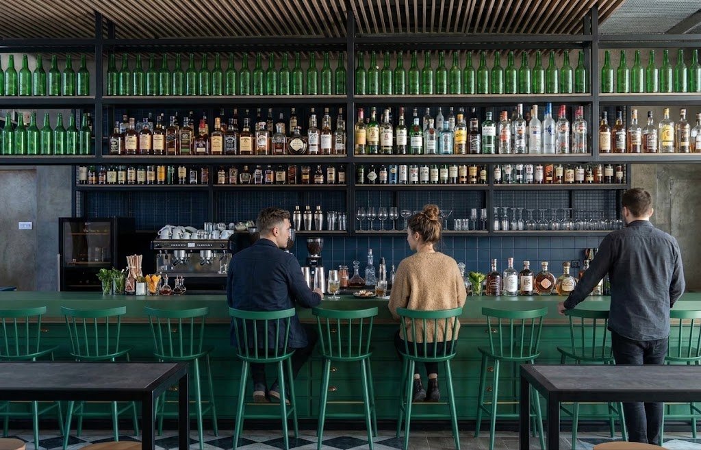 Comptoir de bar moderne vert avec chaises hautes en bois, face à une grande étagère de spiritueux et bouteilles vertes.