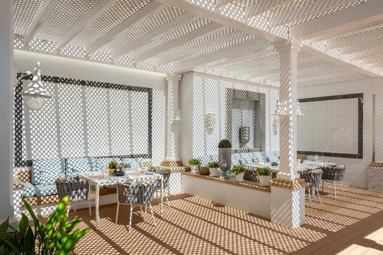 Tables et banquettes élégantes disposées sous une grande pergola blanche en bois sur la terrasse de l'hôtel